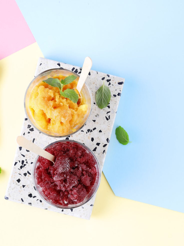 Zwei Gläser mit gelbem und rotem Slush, garniert mit Minze und Holzlöffeln auf pastellfarbenem Hintergrund.