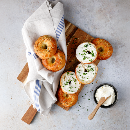 Protein Bagel mit Hanfsamen, Frischkäse und Schnittlauch auf einem Holzbrett angerichtet, daneben liegen weitere Bagels in einem hellen Küchentuch.