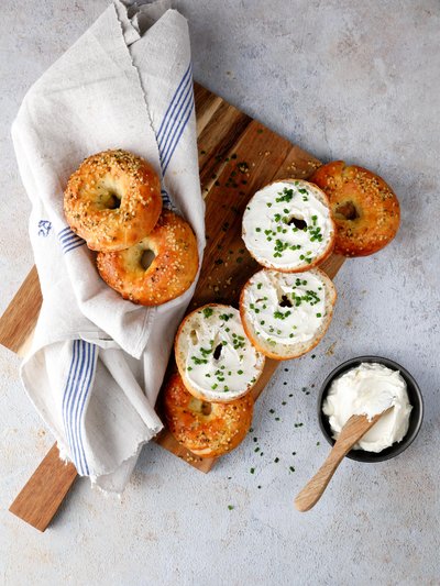 Protein Bagel mit Hanfsamen, Frischkäse und Schnittlauch auf einem Holzbrett angerichtet, daneben liegen weitere Bagels in einem hellen Küchentuch.