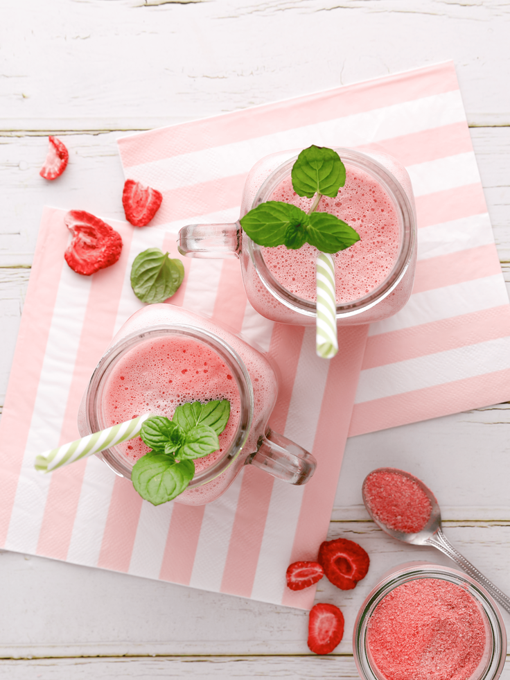 Zwei Gläser mit rosa Erdbeer-Smoothie auf rosa-weiß gestreiften Servietten, jeweils mit grün-weiß gestreiftem Trinkhalm und frischer Minze garniert; daneben liegen getrocknete Erdbeerstücke, ein Glas mit Erdbeerpulver und ein Löffel auf einer weißen Holzoberfläche.