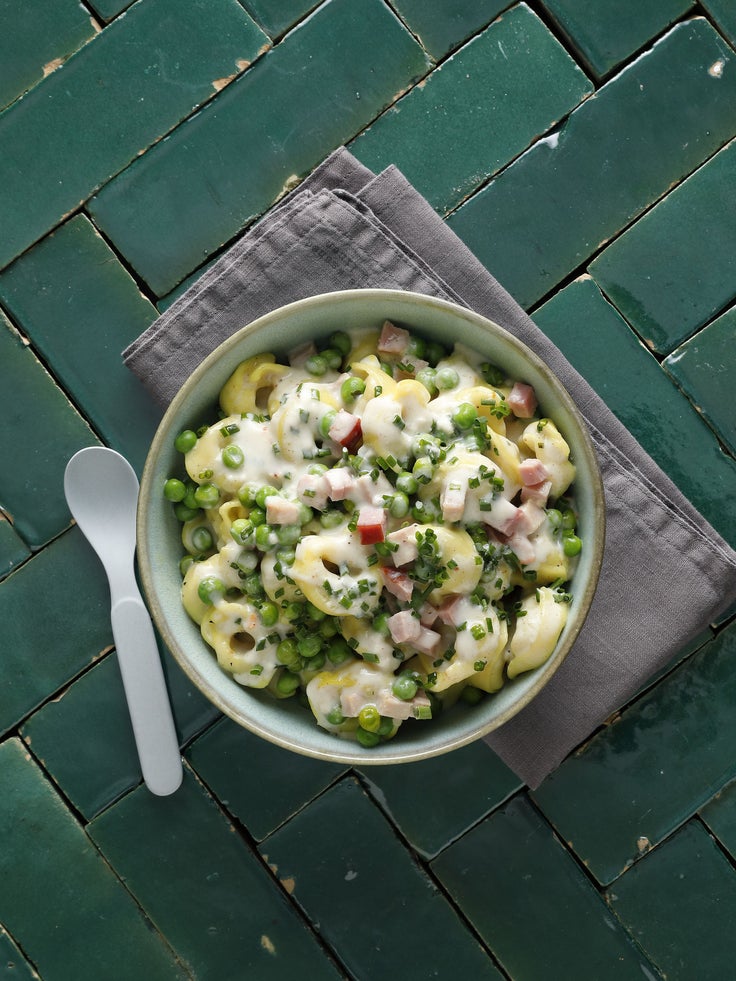 Eine Keramikschüssel gefüllt mit Tortellini, Erbsen und heller Sauce auf einem dunkelgrünen Fliesenspiegel stehend