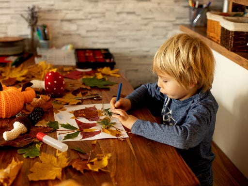 Kleinkind malt konzentriert mit blauem Marker auf weißes Papier, umgeben von Herbst-Naturmaterialien wie Tannenzapfen, bunten Blättern und einem Stoff-Kürbis auf einem Holztisch