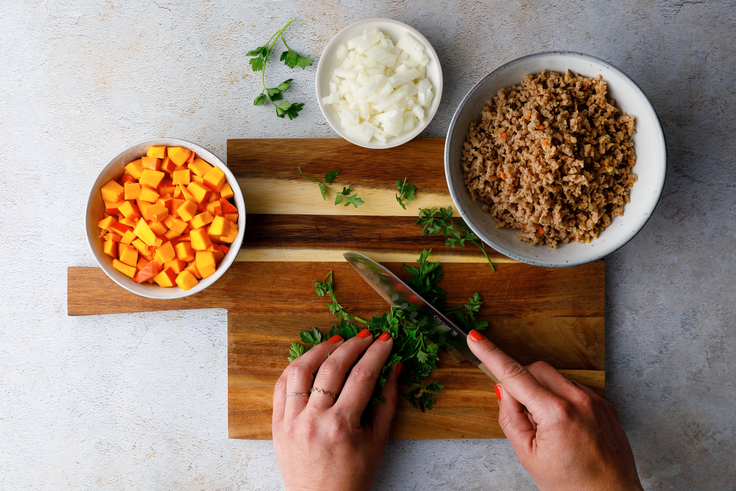 Petersilie wird für Protein Linsen-Spirelli mit Bohnensauce auf einem Holzbrett gehackt, daneben stehen gewürfelter Kürbis, Zwiebel und eingeweichtes Veggiehack.