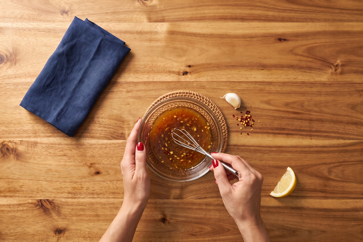 Hände rühren ein würziges Dressing mit Schneebesen in einer Glasschale an, daneben liegen Knoblauch, Chili, Zitrone und ein blaues Küchenhandtuch auf einem Holzhintergrund.