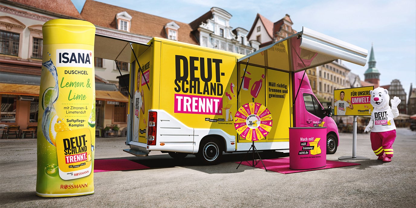 Auf einem Innenstadtplatz steht der gelbe „Deutschland trennt.“-Showtruck mit Glücksrad, Info-Stand und Maskottchen neben einer überdimensionalen ISANA Lemon & Lime Duschgel-Verpackung, um im Rahmen der Mülltrenn-Roadshow von Rossmann und der Initiative „Deutschland trennt.“ für richtige Mülltrennung und Recycling zu werben.