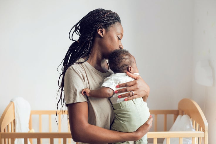 Mutter mit Dreadlocks küsst zärtlich die Stirn ihres Neugeborenen, das sie liebevoll im Arm hält – ein inniger Moment elterlicher Bindung im Kinderzimmer.