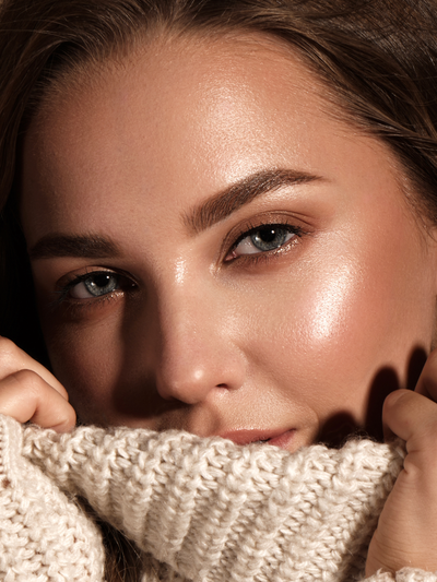Herbst Make-up Essentials: Nahaufnahme einer Frau mit strahlendem Glow-Teint, warmen Bronze-Lidschatten und gepflegten Augenbrauen, die sich in einen cremefarbenen Strickpullover kuschelt