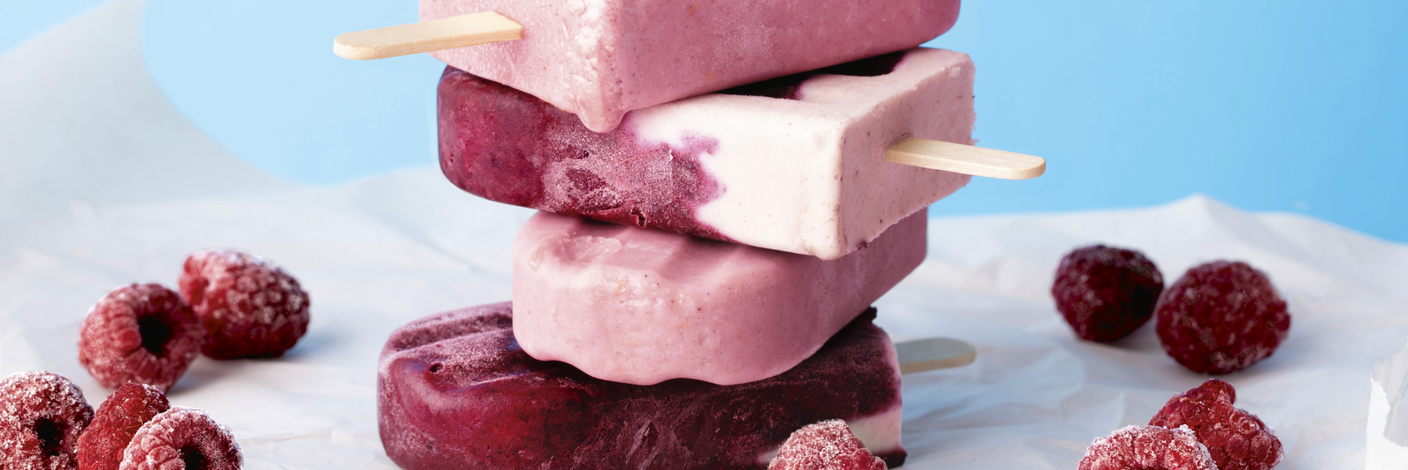 Gestapelte rosa und rote Fruchteis am Stiel mit gefrorenen Himbeeren auf hellem Untergrund vor blauem Hintergrund