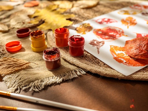 Kind bemalt Herbstblätter mit Pinsel und Gouachefarben in Rot-, Orange- und Gelbtönen auf Jute-Unterlage, um bunte Blattabdrücke auf weißem Papier zu erstellen