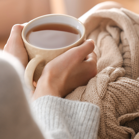 Frau hält eine Tasse heißen Tee in den Händen, eingekuschelt in eine beige Strickdecke für wohlige Wärme und Entspannung.