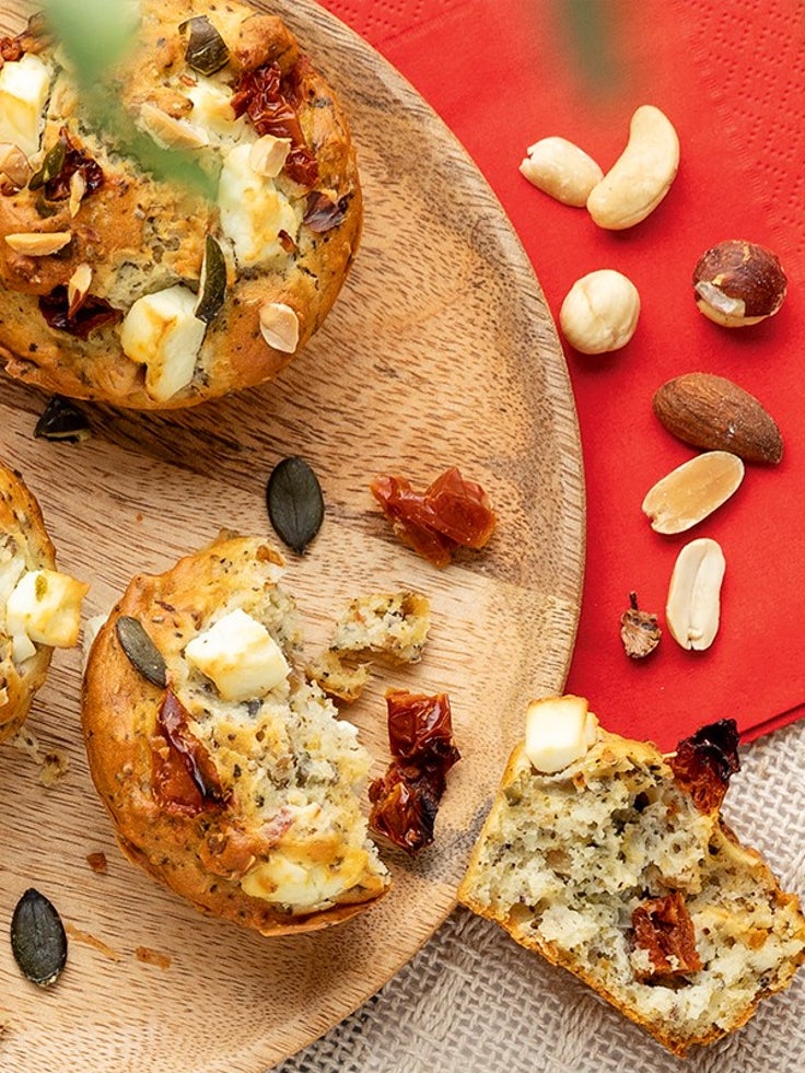 Herzhafte Proteinmuffins mit Feta, Kernen und getrockneten Tomaten auf einem Holzbrett, einer davon aufgebrochen, daneben Nüsse auf einer roten Serviette.