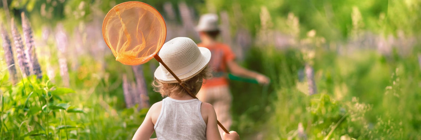 Kleines Kind mit weißem Sonnenhut und Schmetterlingsnetz läuft einen Gartenweg entlang – typische Sommersituation mit erhöhtem Insektenkontakt