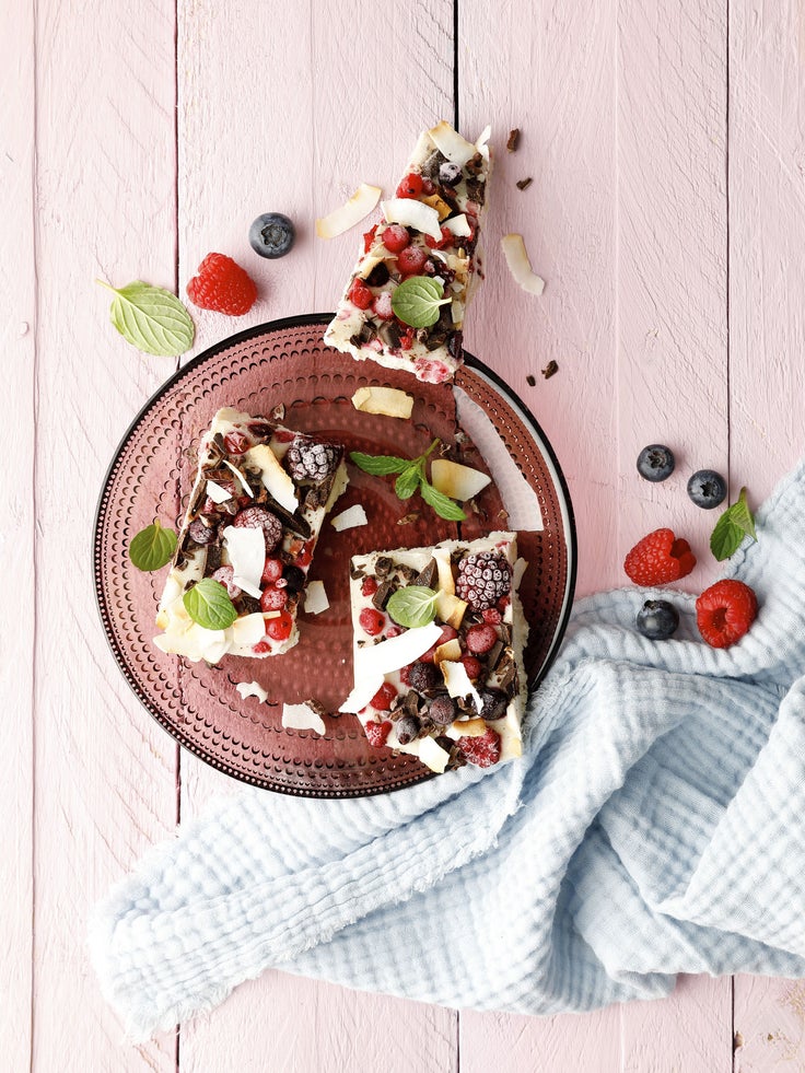 Beerige Froyo Bites auf einem braunen Glasteller, garniert mit Beeren, Kokoschips und Minze auf rosa Holzuntergrund.
