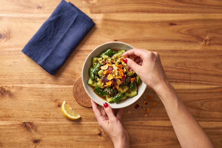 Smashed Cucumber Salat garnieren mit bunten Gemüsechips, Sesam und Dressing, daneben liegen Zitrone und ein blaues Küchenhandtuch auf einem Holzhintergrund.