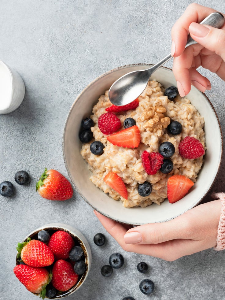 Schüssel mit cremigem Proteinporridge, garniert mit Erdbeeren, Himbeeren, Blaubeeren und Nüssen, eine Hand hält die Schüssel und einen Löffel.