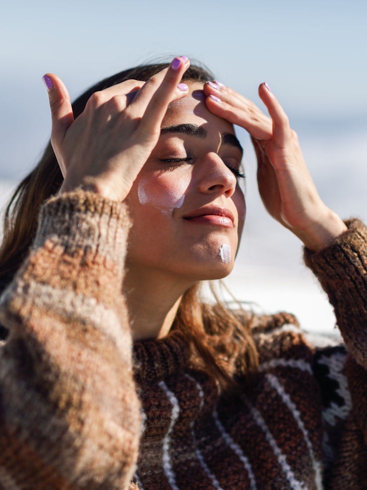 Junge Frau in einem dicken Pullover trägt Sonnencreme im Gesicht auf, um sich vor der Wintersonne zu schützen.
