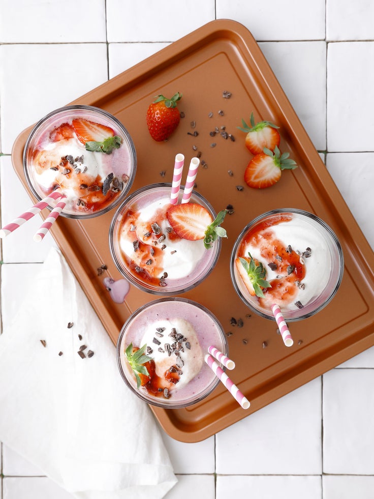 Bildausschnitt eines braunen Tabletts auf weißem Fliesengrund, auf dem vier Milchshakes mit Erdbeeren und rot-weiß-getreiften Strohhalmen stehen, es ist eine Draufsicht .