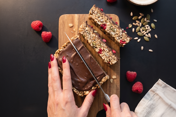 Fruchtige Kokos-Müsliriegel mit Chia-Crunch, Himbeeren und Schokoladenboden werden auf einem Holzbrett in Riegel geschnitten.