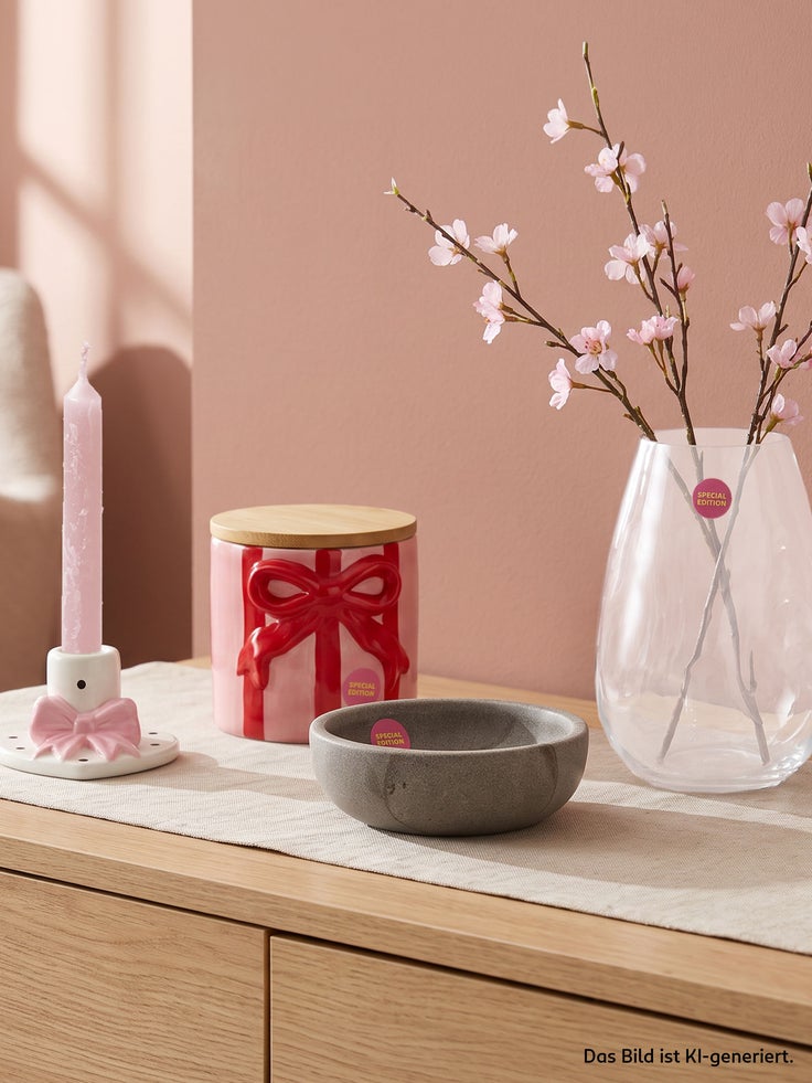 Dekorative Special Edition Wohnaccessoires mit Kerzenhalter, roter Vorratsdose mit Schleife, grauer Schale und Glasvase mit rosa Blüten auf einer Holzkommode