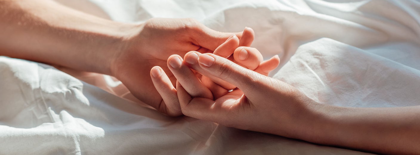 Zwei Hände berühren sich zärtlich auf einem weißen Bettlaken und symbolisieren Intimität sowie Zärtlichkeit im Kontext einer Paarbeziehung nach der Geburt.