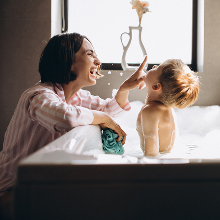 Ein Kind nimmt ein Bad und seine Mutter kniet davor und macht Spaß mit ihm.