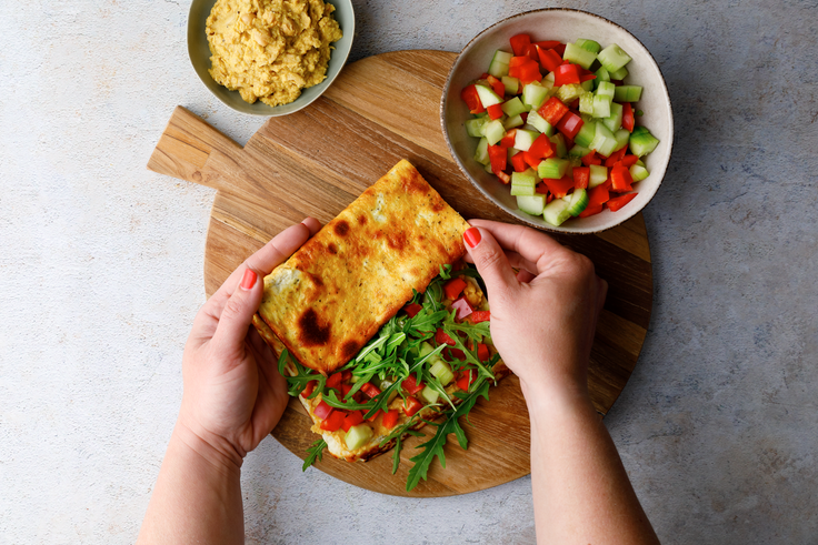 Protein Wrap mit Hummus wird mit Kichererbsenmasse, Gemüsewürfeln und Rucola gefüllt und auf einem Holzbrett zusammengeklappt.