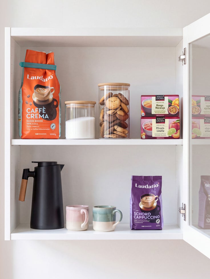 alt="Offener Küchenschrank mit Laudatio Caffè Crema und Schoko Cappuccino, Teeboxen, Keksgläsern, Kaffeekanne und Tassen"