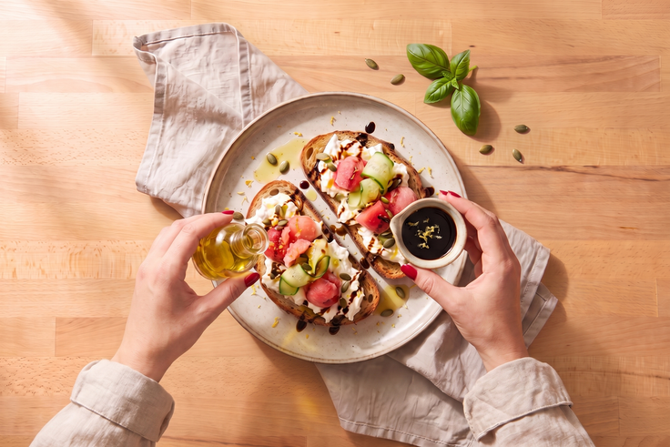 Hände verfeinern Burrata Sommerbrot mit Olivenöl und Balsamico-Creme, garniert mit Melonen, Gurkenstreifen, Kürbiskernen und Zitronenabrieb auf einem Holzhintergrund.