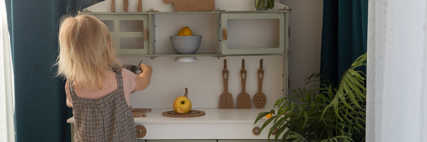 Kleinkind spielt an einer Kinderküche mit Holzspielzeug und Obst in einem hellen Wohnraum
