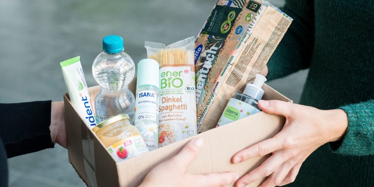 Auf der Grafik ist eine Kiste mit mehreren Drogerieartikeln zu sehen, die von zwei Personen gehalten wird