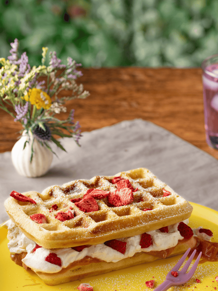 Ein Holztisch mit einer Leinen Tischdecke, auf dem ein gelber Teller mit einem Waffelkuchen und einem Wiesenstrauch stehen