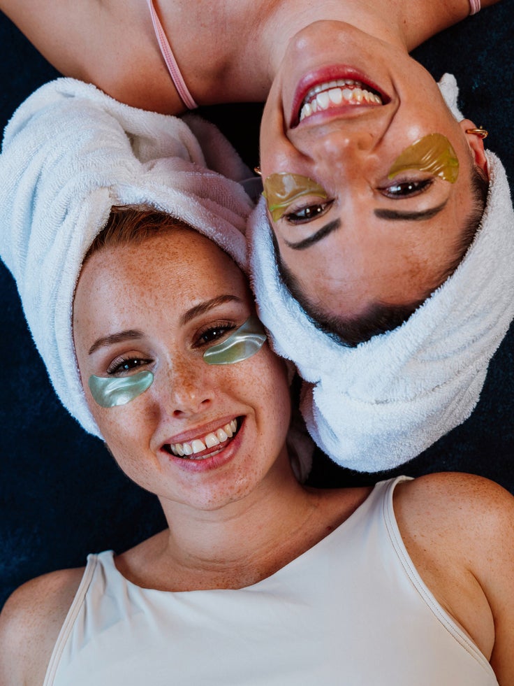 Zwei Frauen, die lachend mit Augenpads und einem Haarturban auf dem Boden liegen