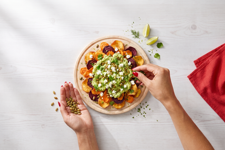 Schritt 3: Loaded Gemüse Chips werden mit Avocadocreme, Feta, Kräutern und Kürbiskernen auf einem Holzteller garniert.