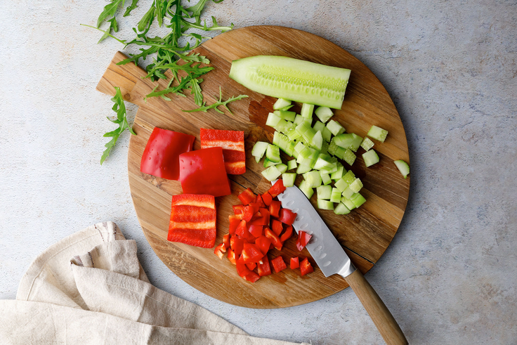 Paprika und Gurke werden für Protein Wraps mit Hummus auf einem Holzbrett gewürfelt, daneben liegt Rucola auf heller Arbeitsfläche.