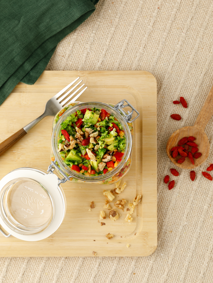 Bildausschnitt von oben auf ein Holzbrett mit einem geöffneten Einwegglas gefüllt mit einem bunten Salat, daneben liegt eine Gabel links  und rechts Gojibeeren