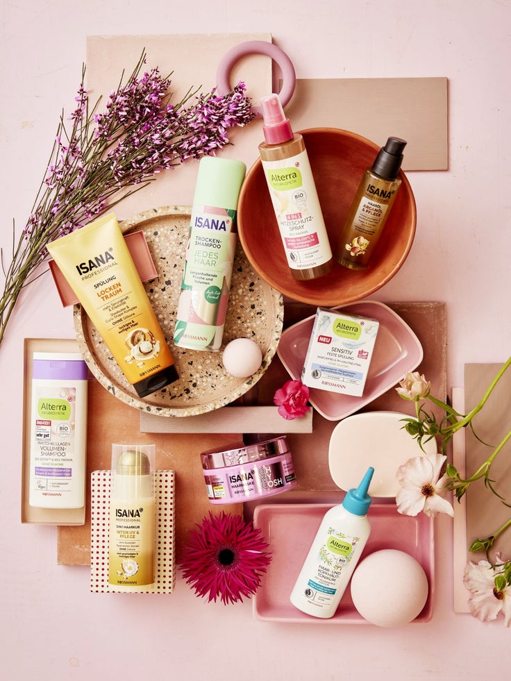 Dekoratives Flatlay mit ISANA und Alterra Haarpflegeprodukten wie Shampoo, Spülung, Hitzeschutz, Haaröl und Haarkur auf rosa Hintergrund mit Blumen und Keramikschalen