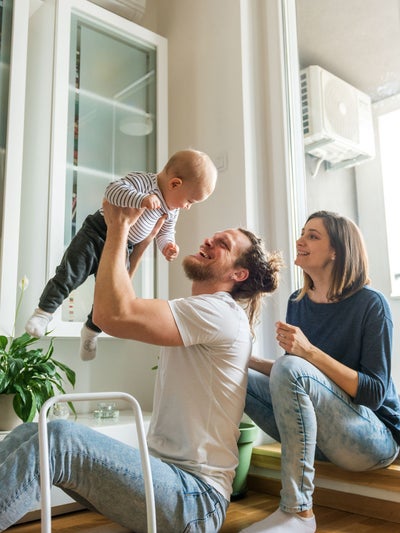 Vater hebt lachend sein Baby in die Luft, während die Mutter daneben auf der Fensterbank sitzt und zuschaut