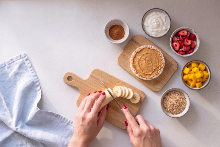 Banane wird für Reiswaffel Variationen in Scheiben geschnitten, daneben stehen Joghurt, Erdbeeren, Mango, Sesam und eine Reiswaffel mit Nussmus.