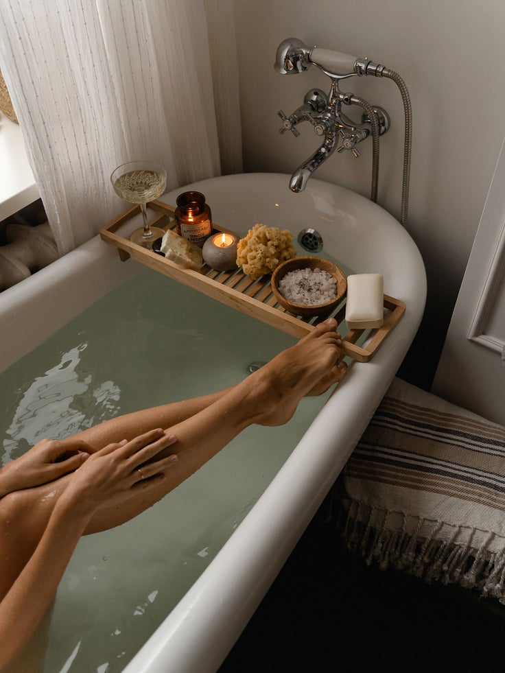 Eine mit Wasser gefüllte Badewanne von oben mit zwei Frauenbeinen im Blickfeld und Badeprodukten auf dem Badewannenrand