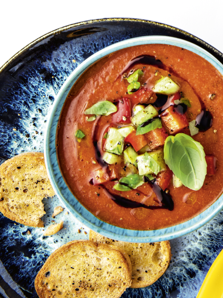 Tomaten-Gazpacho mit Gurken, Paprika, Basilikum und Balsamico-Topping, serviert mit gerösteten Brotscheiben.