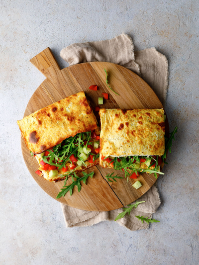 Protein Wraps mit Hummus, Rucola, Gurke und Paprika auf einem runden Holzbrett angerichtet.