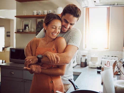 Ein glückliches Paar umarmt sich lachend in der Küche und symbolisiert Freude und Verbundenheit in der Partnerschaft nach der Geburt.