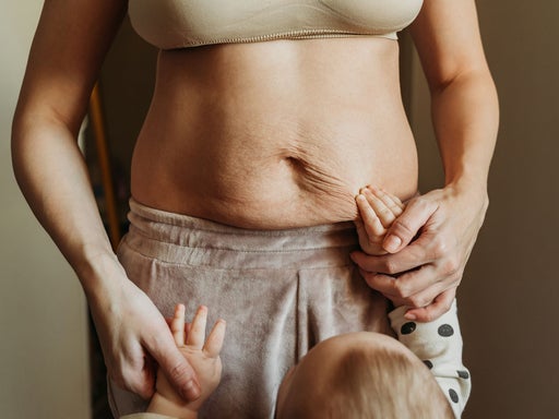 Eine Mutter hält ihr Baby, das ihren Bauch berührt und die körperlichen Veränderungen nach der Geburt, wie Dehnungsstreifen, sichtbar macht.