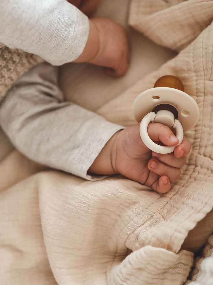 Baby liegt unter einer weichen Decke und hält einen Schnuller mit Ringgriff in der Hand.