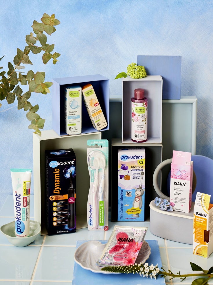 Rossmann Pflege- und Dentalprodukte von Alterra, Prokudent und ISANA in pastellfarbenen Regalfächern vor hellblauem Hintergrund mit Pflanzen und Blumen