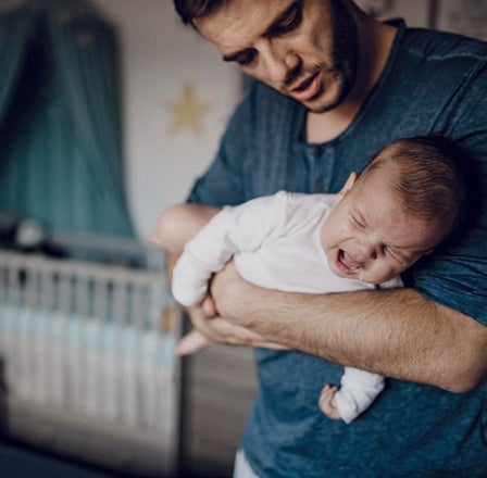 Vater hält ein weinendes, strampelndes Baby im Kinderzimmer, das zur Schlafenszeit nicht einschlafen möchte"