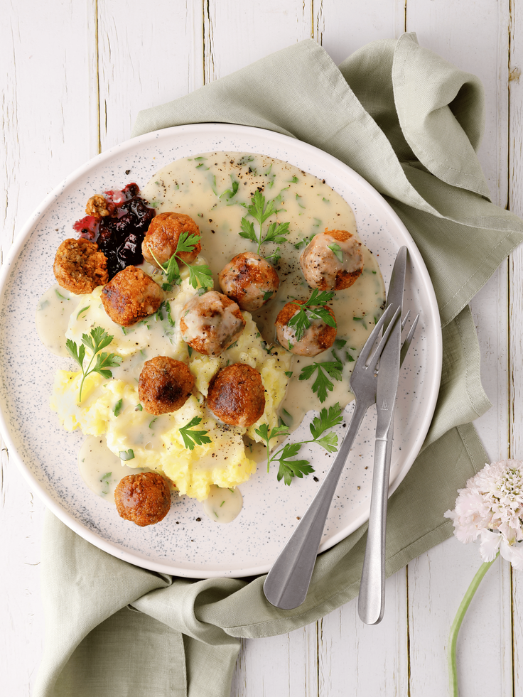 Teller mit veganen Köttbullar, Kartoffelstampf, cremiger Soße und Petersilie, angerichtet mit Besteck auf hellem Holztisch.
