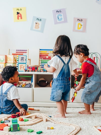 Drei Kleinkinder spielen gemeinsam in einem hellen Spielzimmer, das mit pädagogischem Spielzeug wie einer Holzeisenbahn, einem Abakus und Alphabet-Karten ausgestattet ist.
