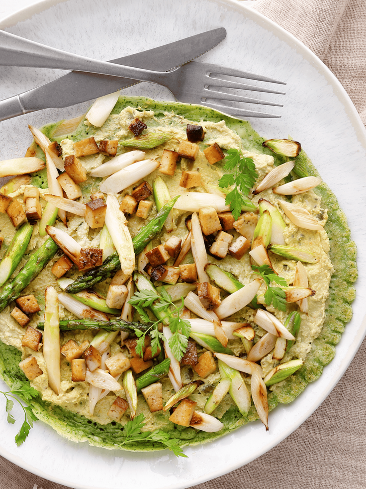  Grüner Pfannkuchen auf einem weißen Teller, belegt mit angebratenem Spargel, gewürfeltem Tofu und frischen Kräutern, daneben Messer und Gabel auf einer beigefarbenen Serviette.