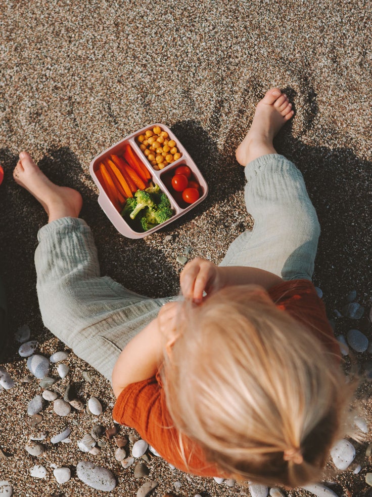 Ein Kind von oben fotografiert sitzt im Sand mit Muscheln ringsherum, vor ihm steht eine Snackbox